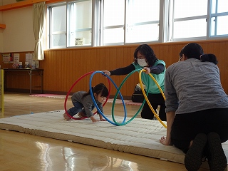 白いマットの上で、二人の大人が支えるフープを、小さな女の子がしゃがんでくぐっている写真