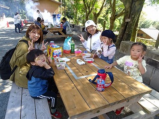 屋外のピクニックテーブルを囲んで座る親子2組が、テーブルにお弁当を広げ、みんなでポーズをとっている写真