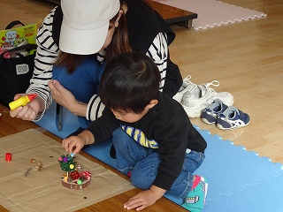 室内のマットの上で、木の実や木の枝などをボンドで貼り付けながらクリスマスの飾りを作っている親子の写真