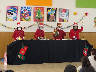 靴下やサンタクロースの装飾がされた室内で、赤い衣装にサンタ帽を被った4人の女性が、ハンドベルの演奏をしている写真