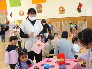 クリスマスの装飾がされた室内で、大人や子供達が見守る中、子供達が楽しそうにおもちゃのハンマーを握って手作りのもぐらたたきで遊んでいる様子の写真