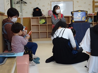 女性が紙芝居の読み聞かせてしており、親子連れの家族らが床に座り、話を聞いている様子の写真