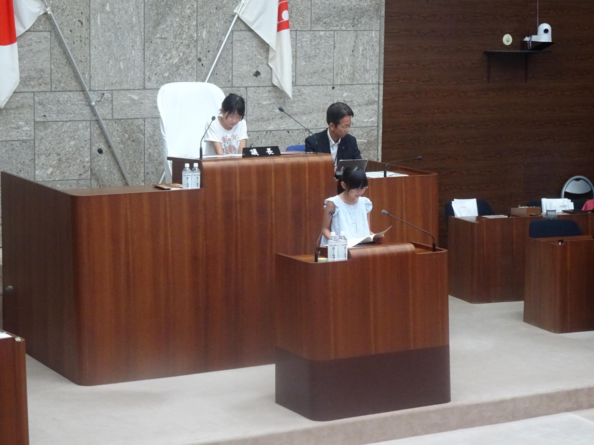 市議会議場で、議長席に小学生の女の子と議員席に男性が座り、その前の演壇に立って資料を読んでいる小学生の女の子の写真