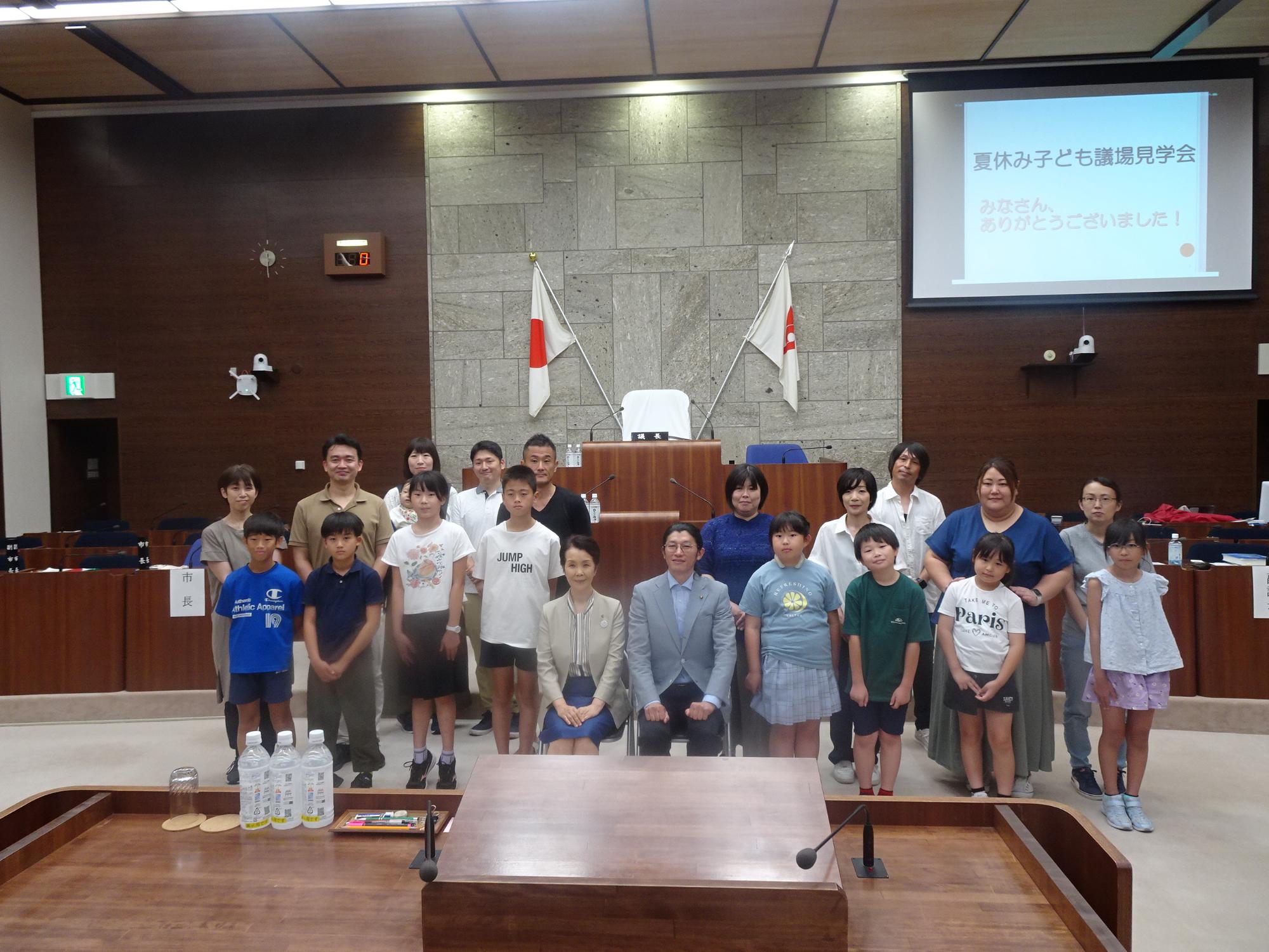 市議会議場で、お仕事体験を行った小学生達と保護者、付き添いの参加者が「夏休み子ども議場見学会」の看板を背景に記念撮影している写真