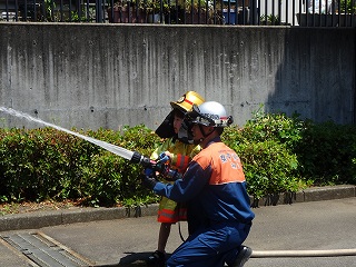 屋外で、消防士の服装をした5歳くらいの子供が消防士と一緒にホースを支え、勢いよく水を放水している消防体験の写真
