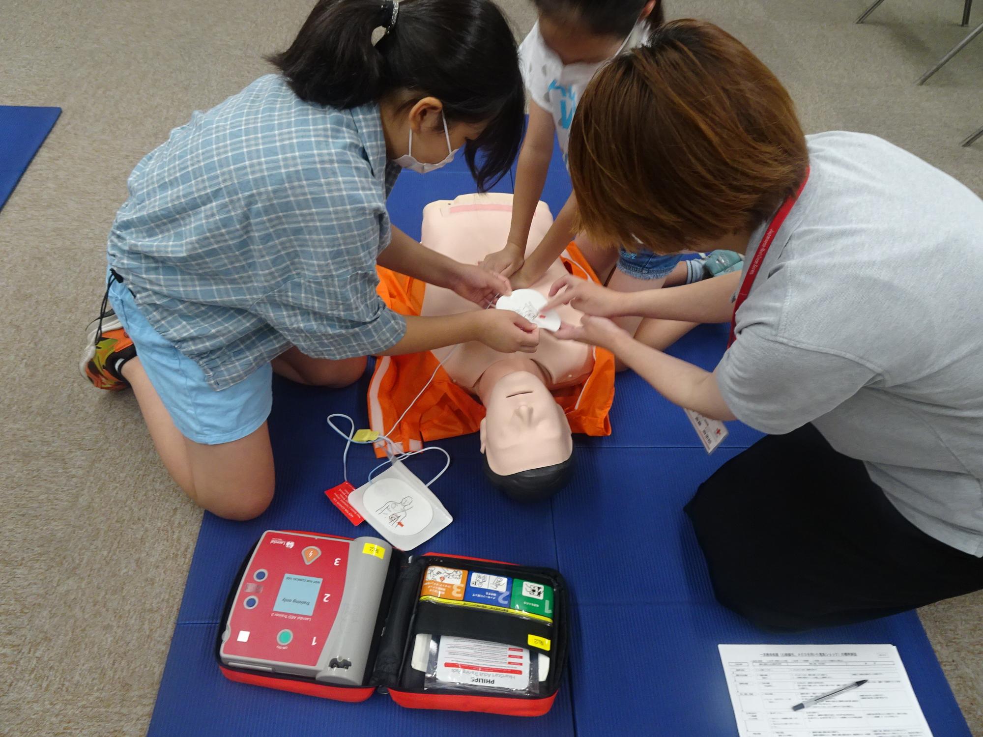 二人の子供と大人が一緒に、床に置かれたマネキンの胸にAEDの電極パッドを張り付け、機材の使い方を体験している写真