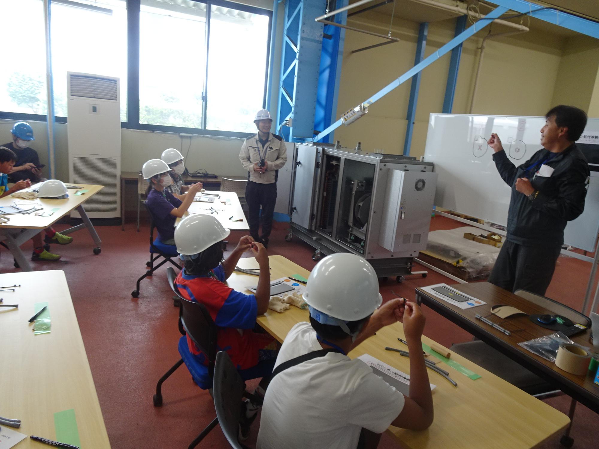 広い室内で、ヘルメットを被った子供達が席に座っており、ホワイトボードを使って説明する男性の話を聞いている写真