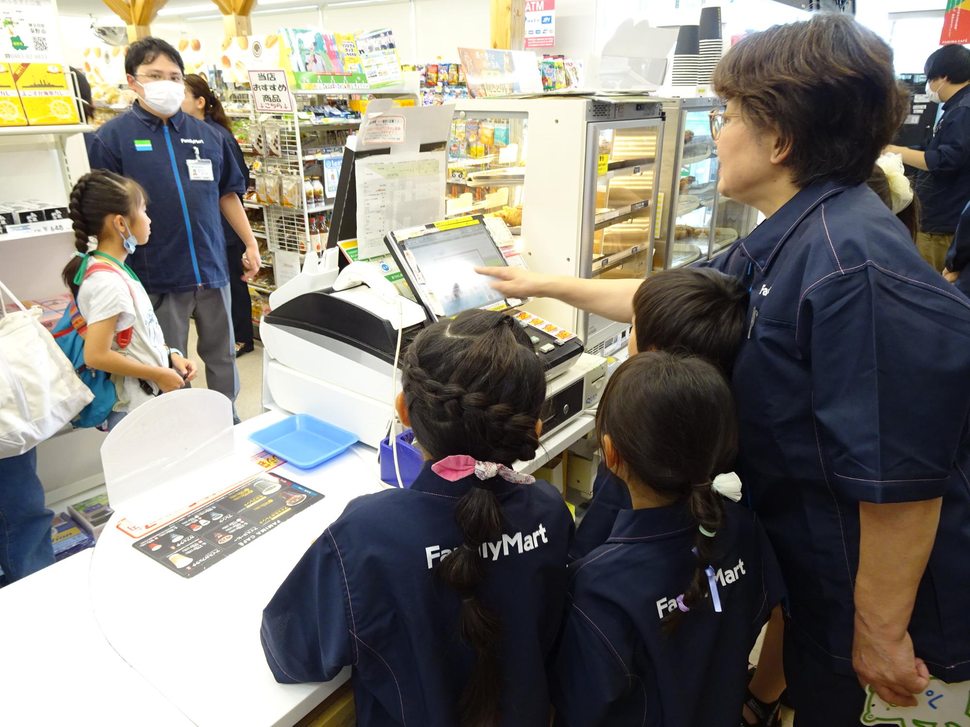 紺色のユニフォームを着用した子供達が、店員にレジの使い方を教わりながら、コンビニエンスストアで職業体験をしている写真