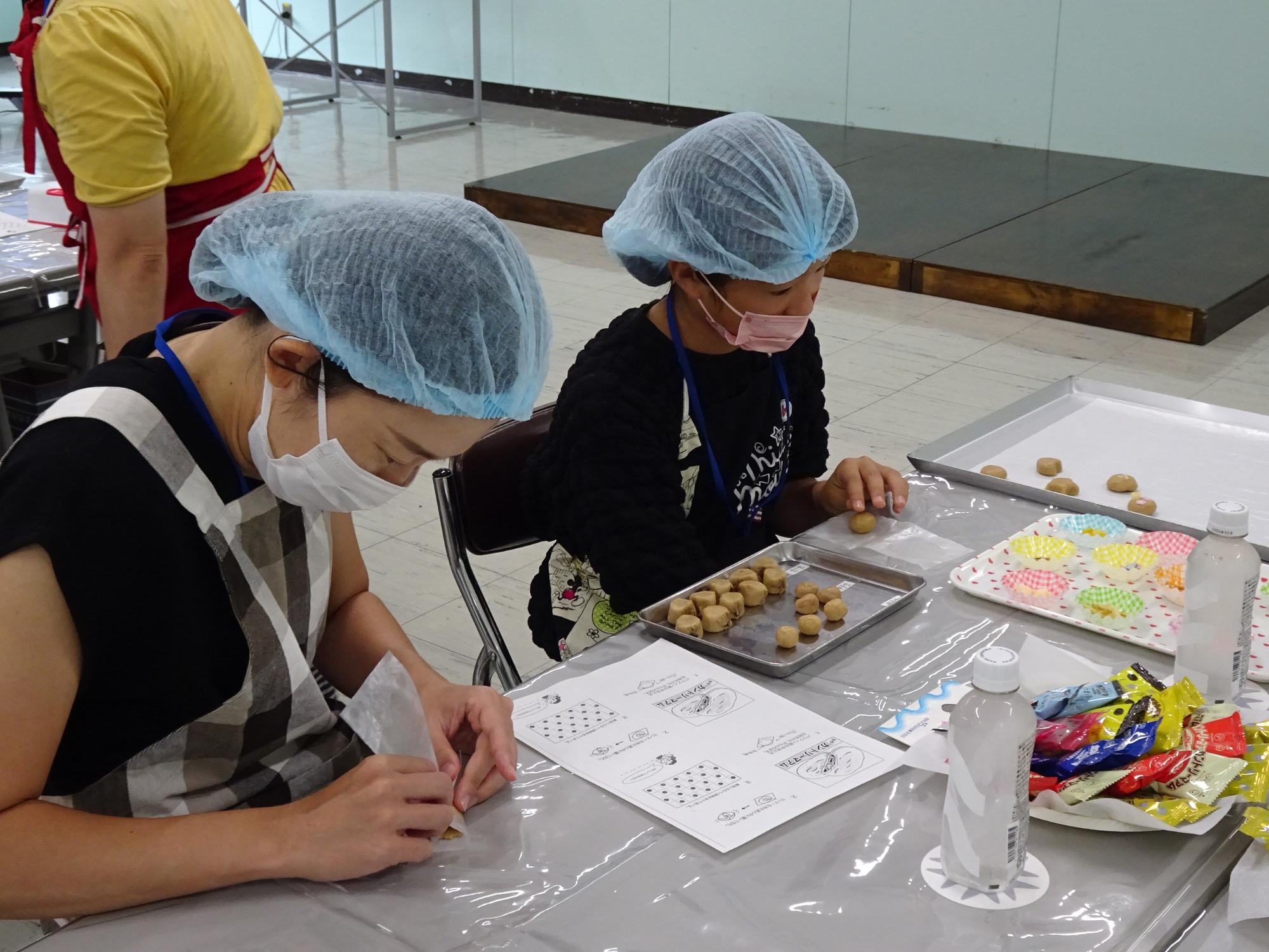 エプロンにマスク、ヘアキャップを着用した親子が、テーブル上のトレーに並べてお菓子作りに取り組んでいる様子の写真