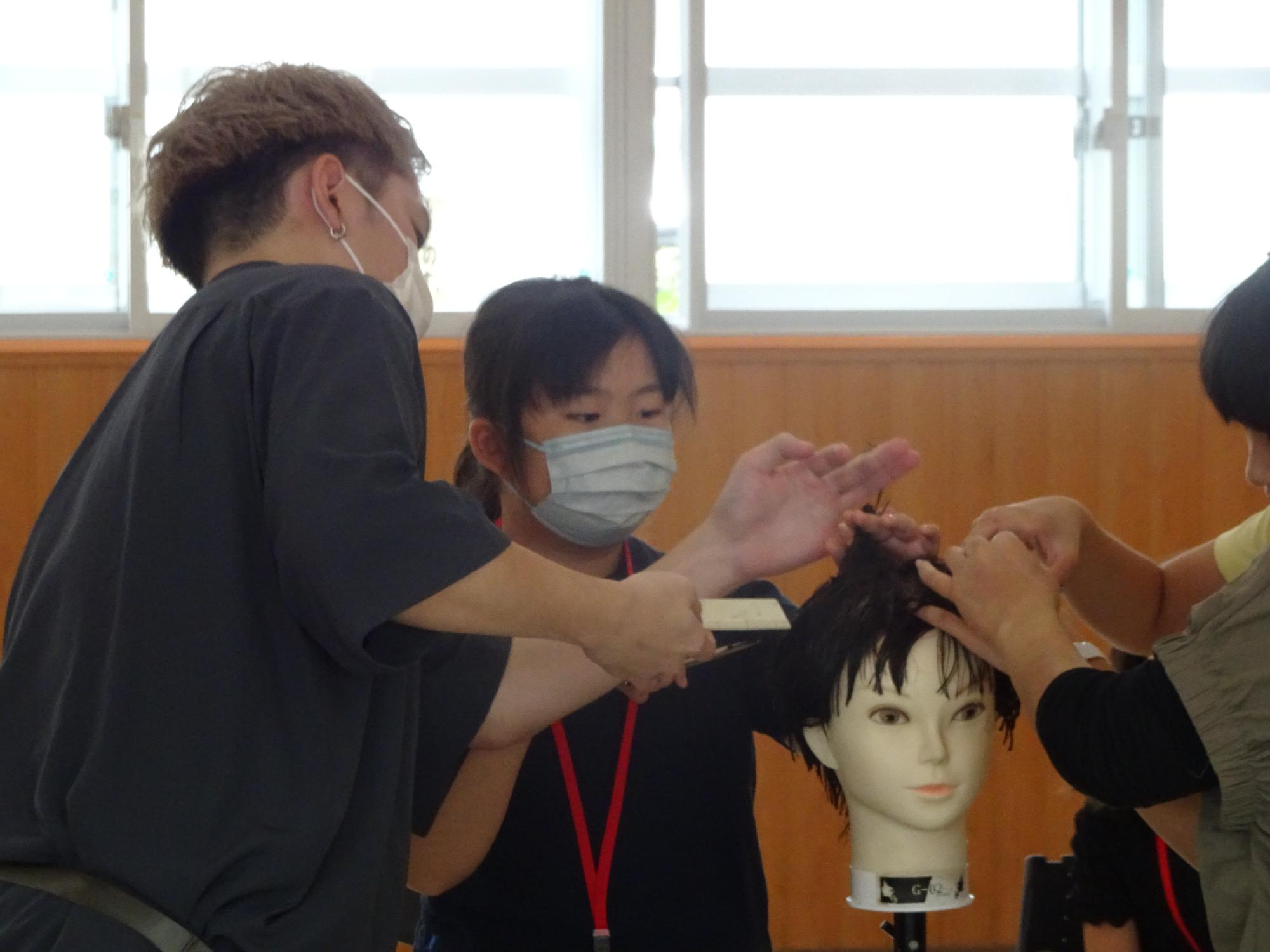 美容師の男性が、小学生の女の子にマネキンを使って髪の毛のカットの仕方を教えている写真