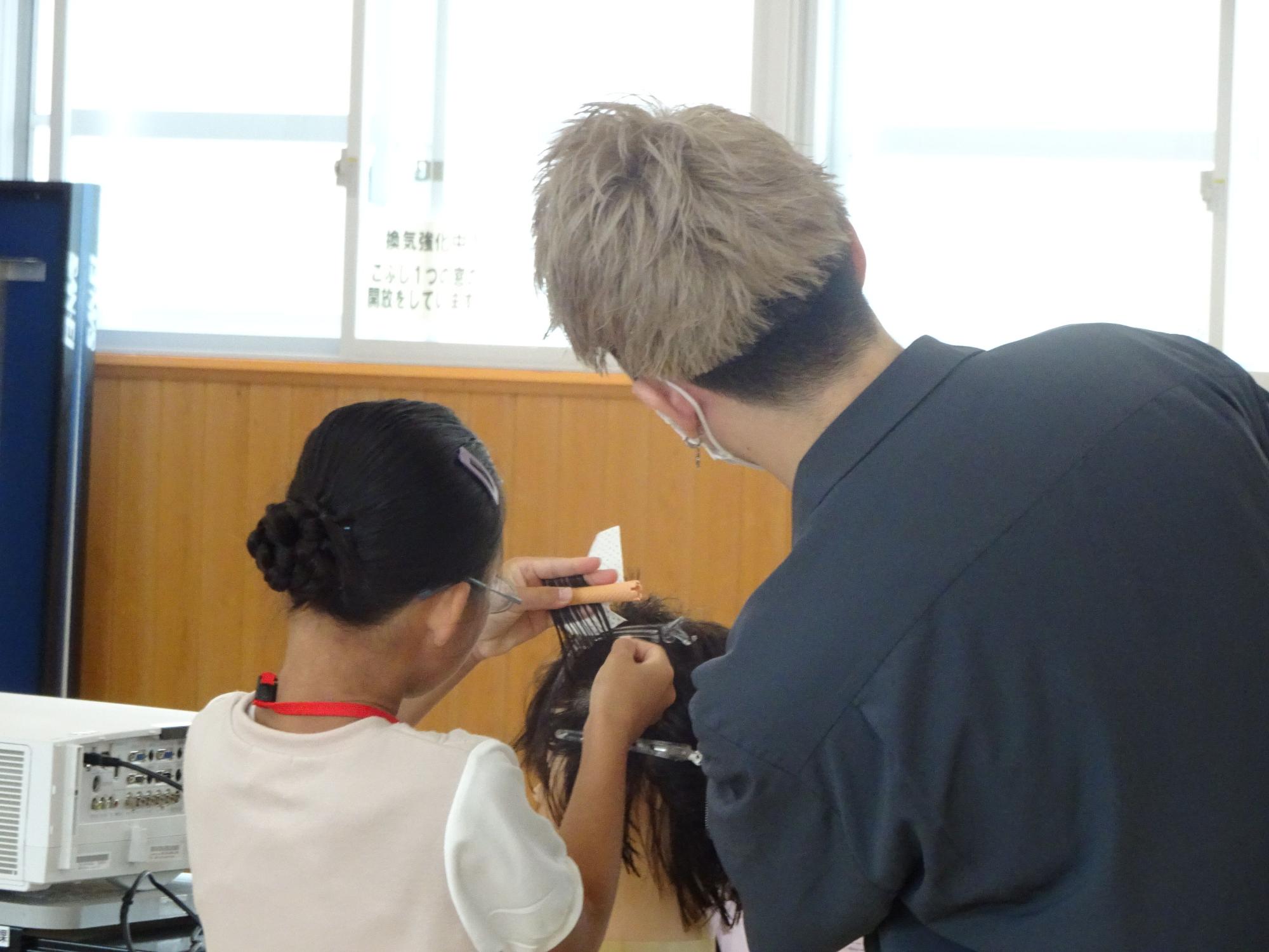 美容師の男性が、小学生の女の子にマネキンを使って髪をロッドに巻き付ける作業を教えている写真
