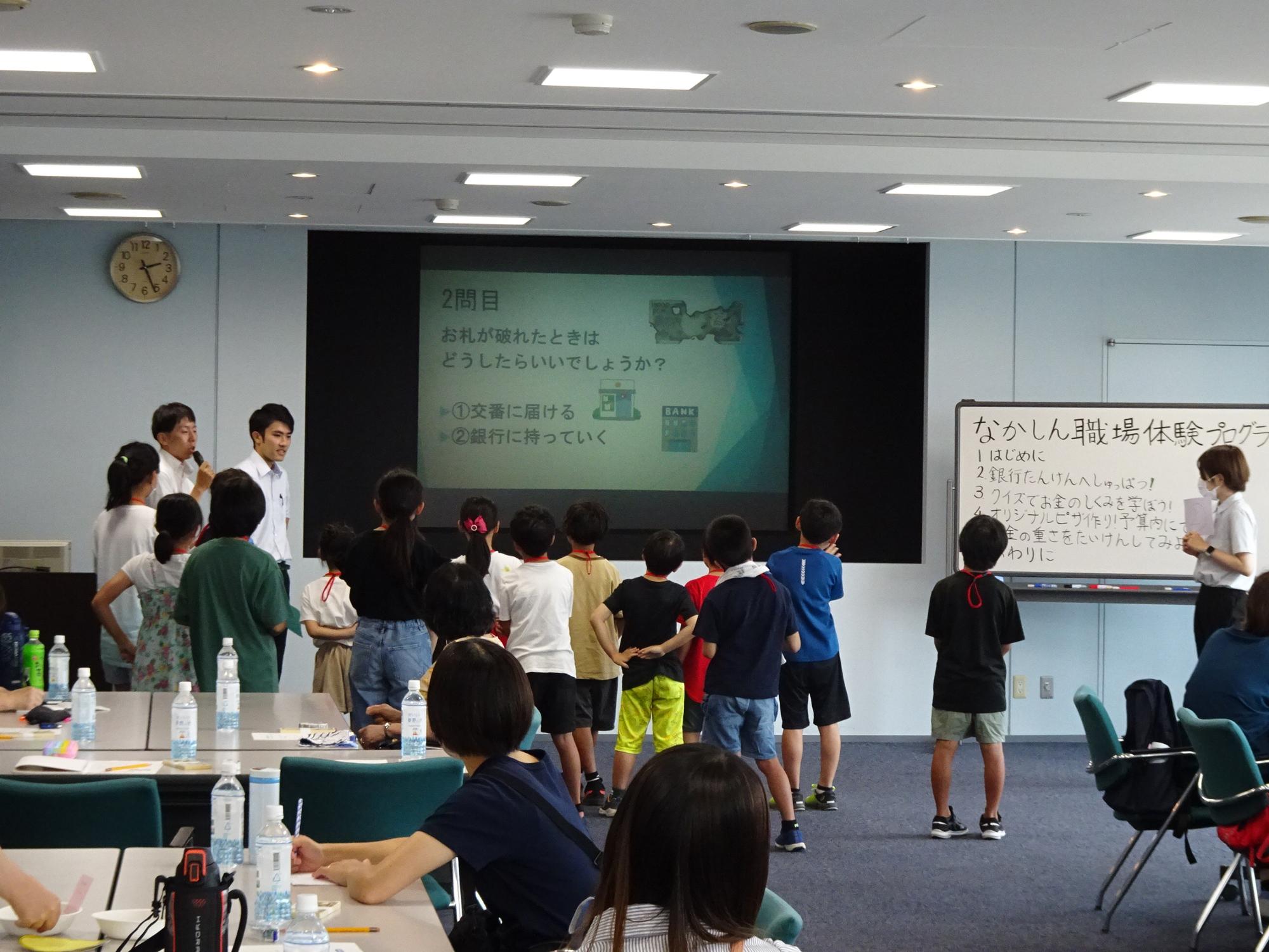 保護者、付き添いの参加者は席に座り、小学生達はプロジェクターに映し出された資料の前に立ち、金融機関の職員の話に耳を傾けている写真