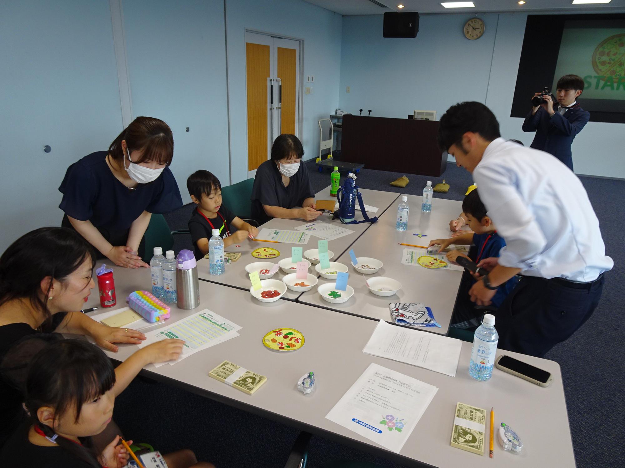子供達と保護者、付き添いの参加者がテーブルを囲み、複数の皿にカラフルなお金のようなものが並べられ、職員の説明に耳を傾ける様子の写真