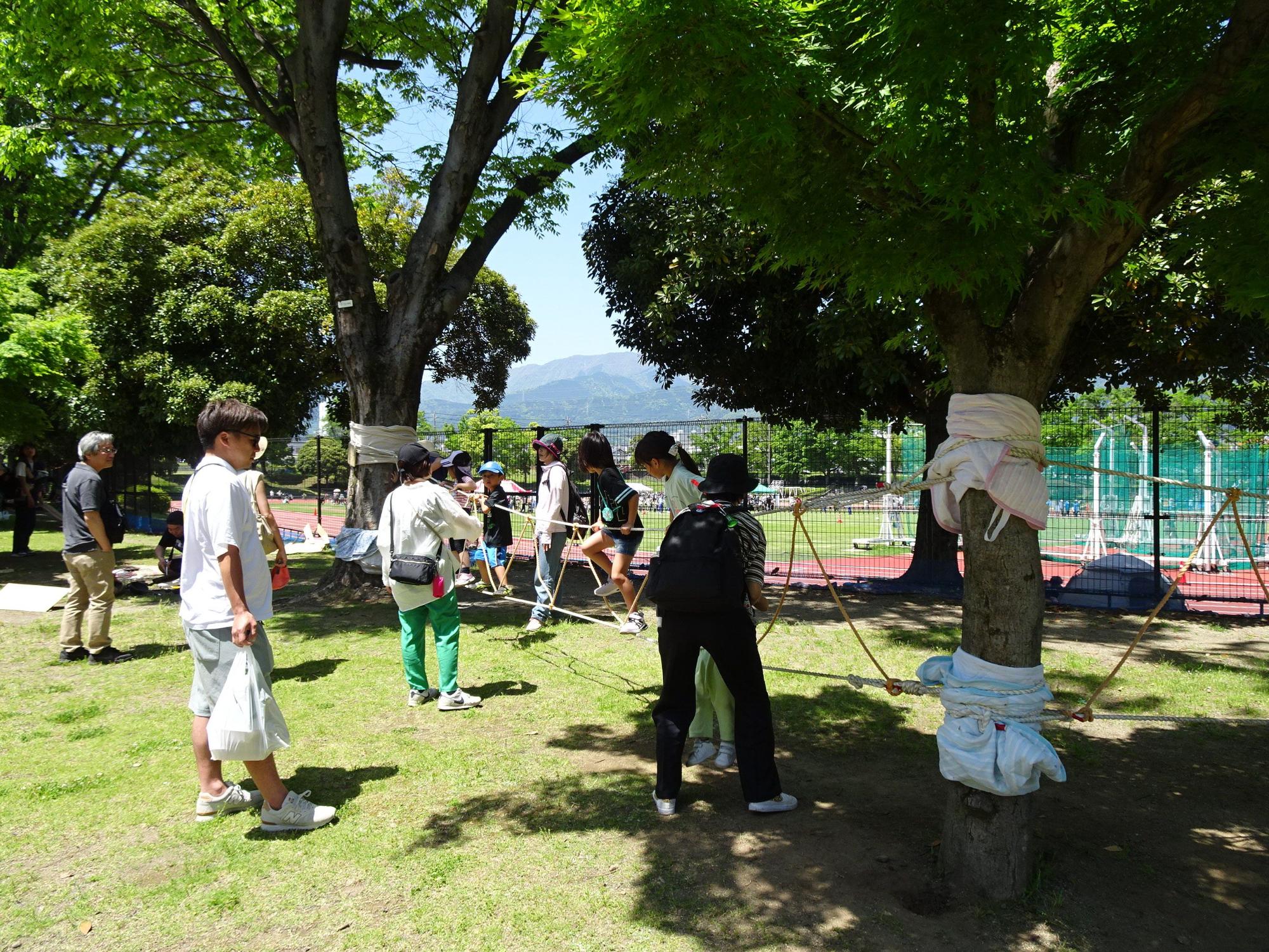 晴れた公園の木陰の下の芝生で、数人の子どもたちが、木の幹に巻き付けられたロープをジャンプしたり跨いだりして進むアスレチックのような活動を、数人の大人たちが見守っているイベント風景の写真