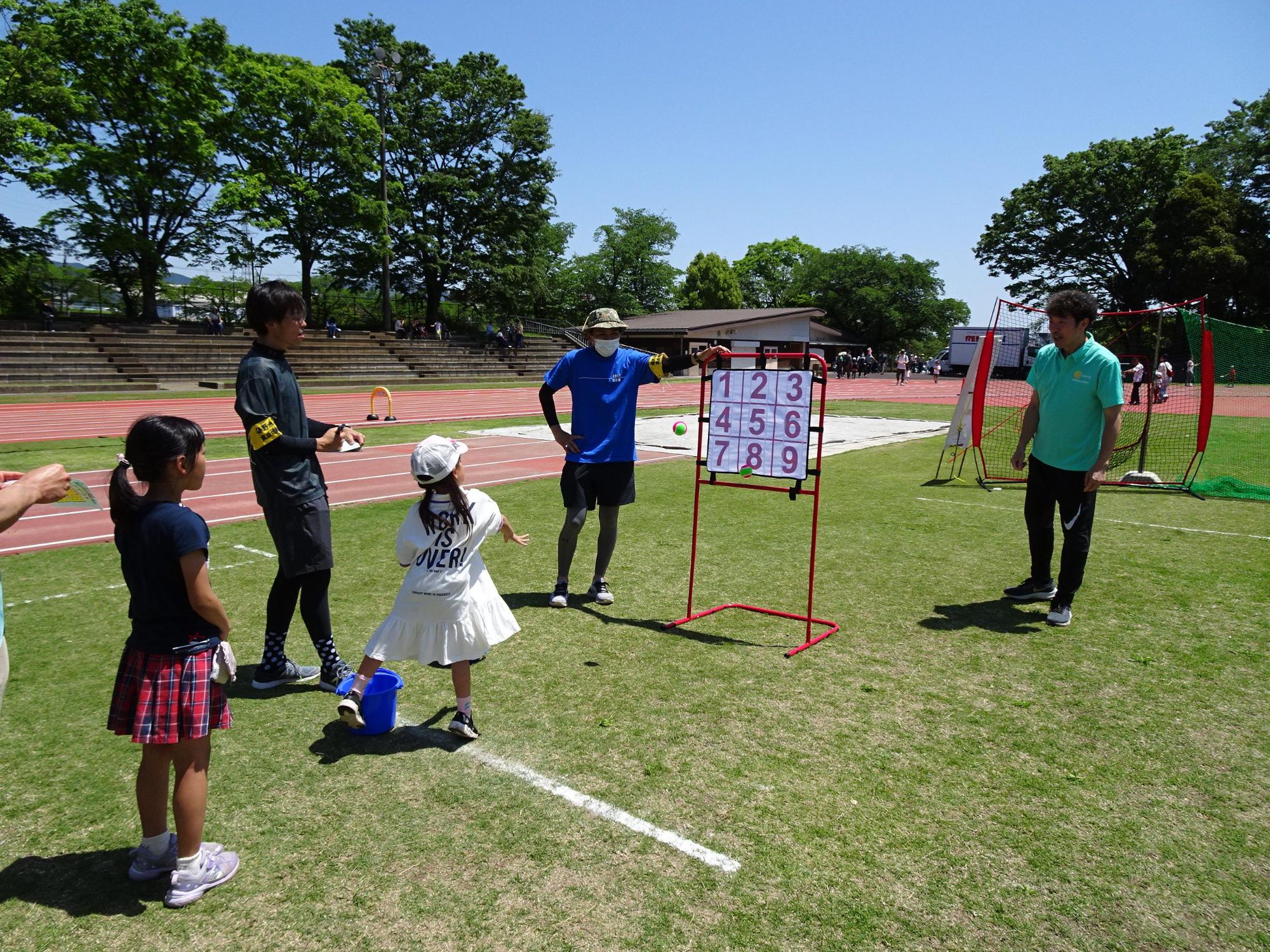 晴天の屋外競技場で、芝生の上に置かれた九つの数字が書かれたストラックアウトの的をめがけて、白いワンピースの少女がボールを投げようとしている様子を、二人の指導員と一人の少女が見守っている写真