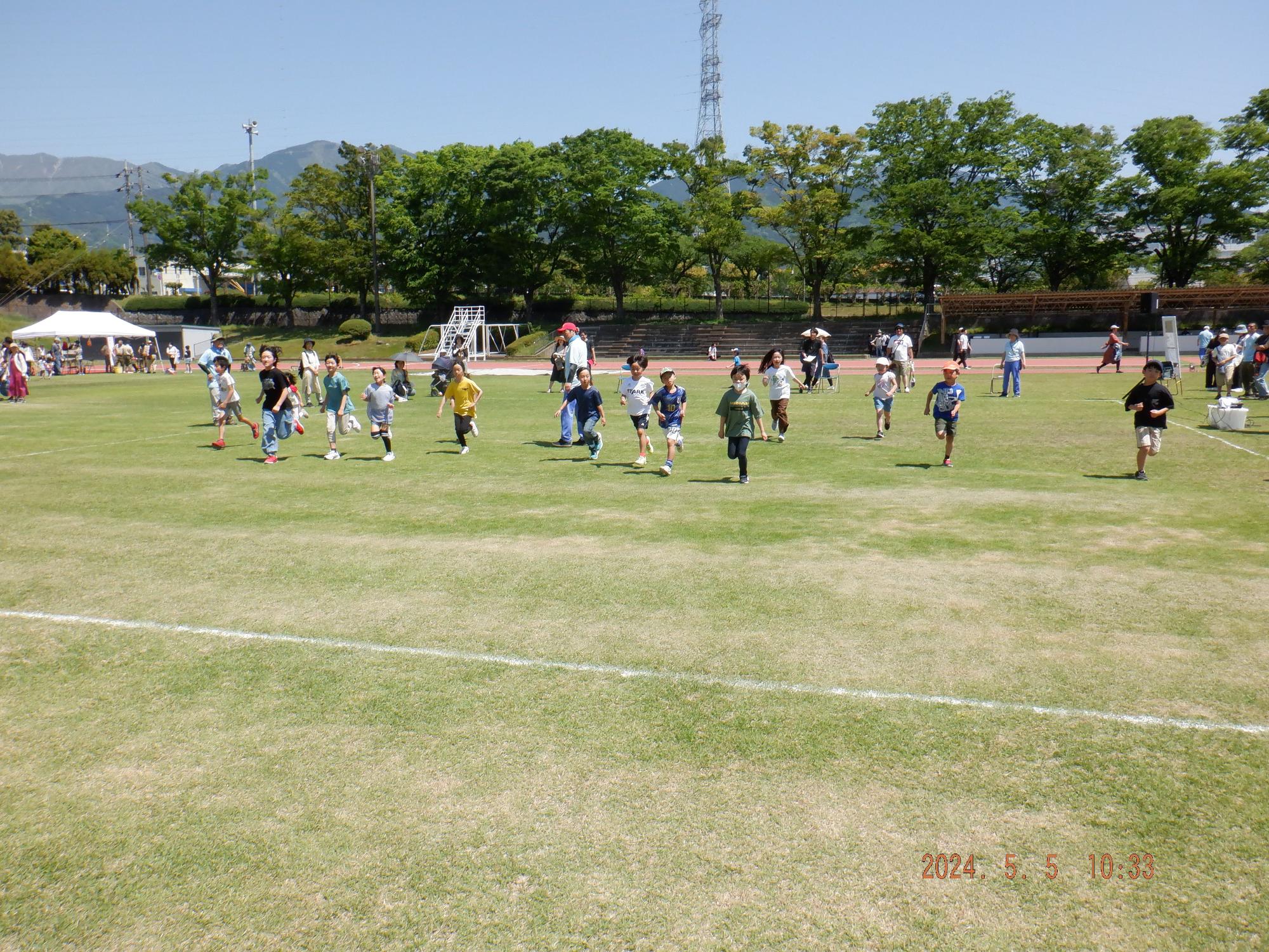 晴天の運動場で、芝生の広場の中央を様々な服装をした大勢の子どもたちが一斉に駆け抜けている様子を、トラックや観客席の周辺にいる数人の大人が見守っている、イベント風景の写真
