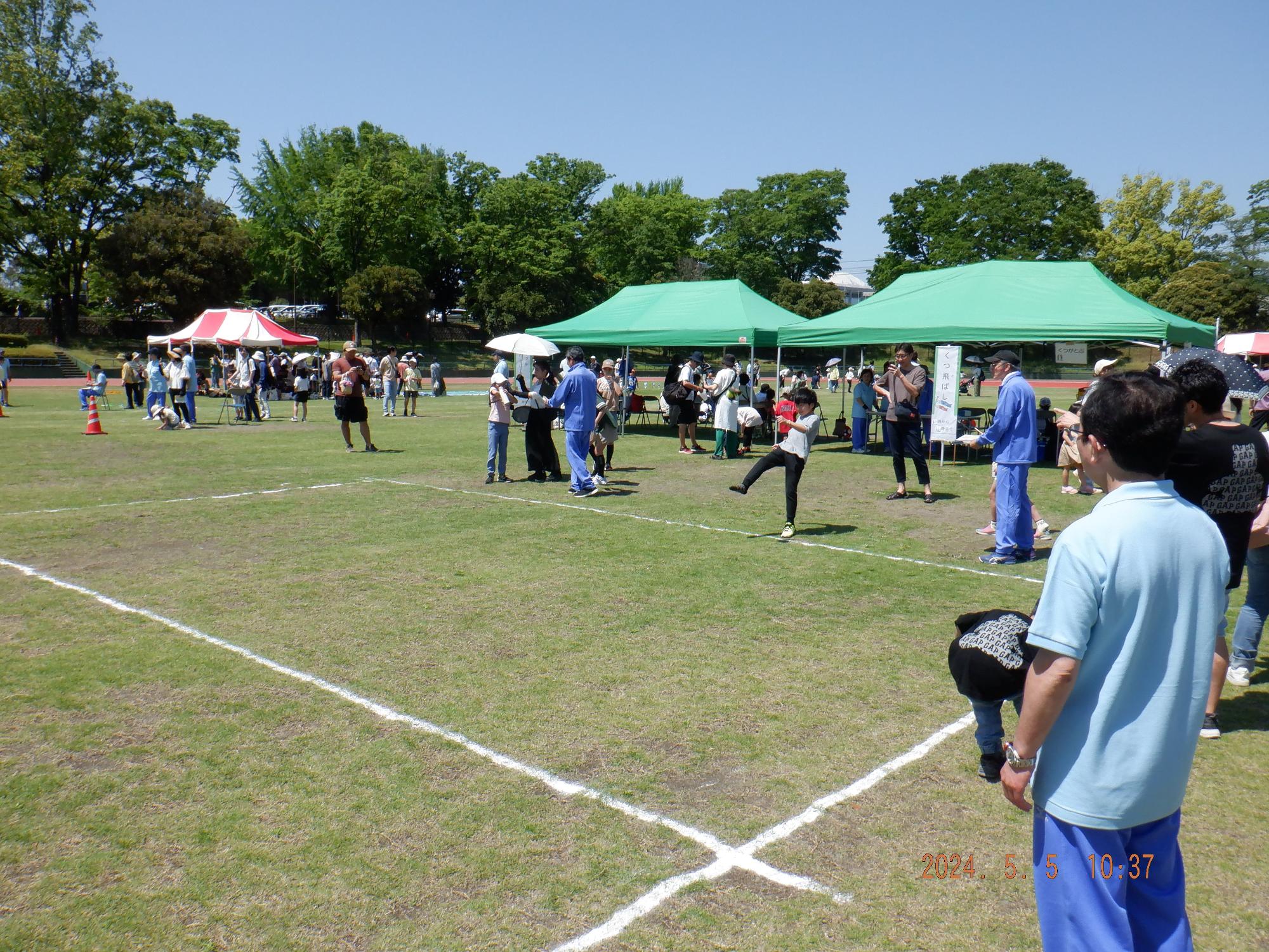 晴れた屋外の芝生広場で、中央に白い線で区切られたエリアがあり、その周囲に緑や赤のテントが設置され、多くの人々が立ち話やイベントに参加している中、一人の少年がボールを蹴るようなポーズをしている写真