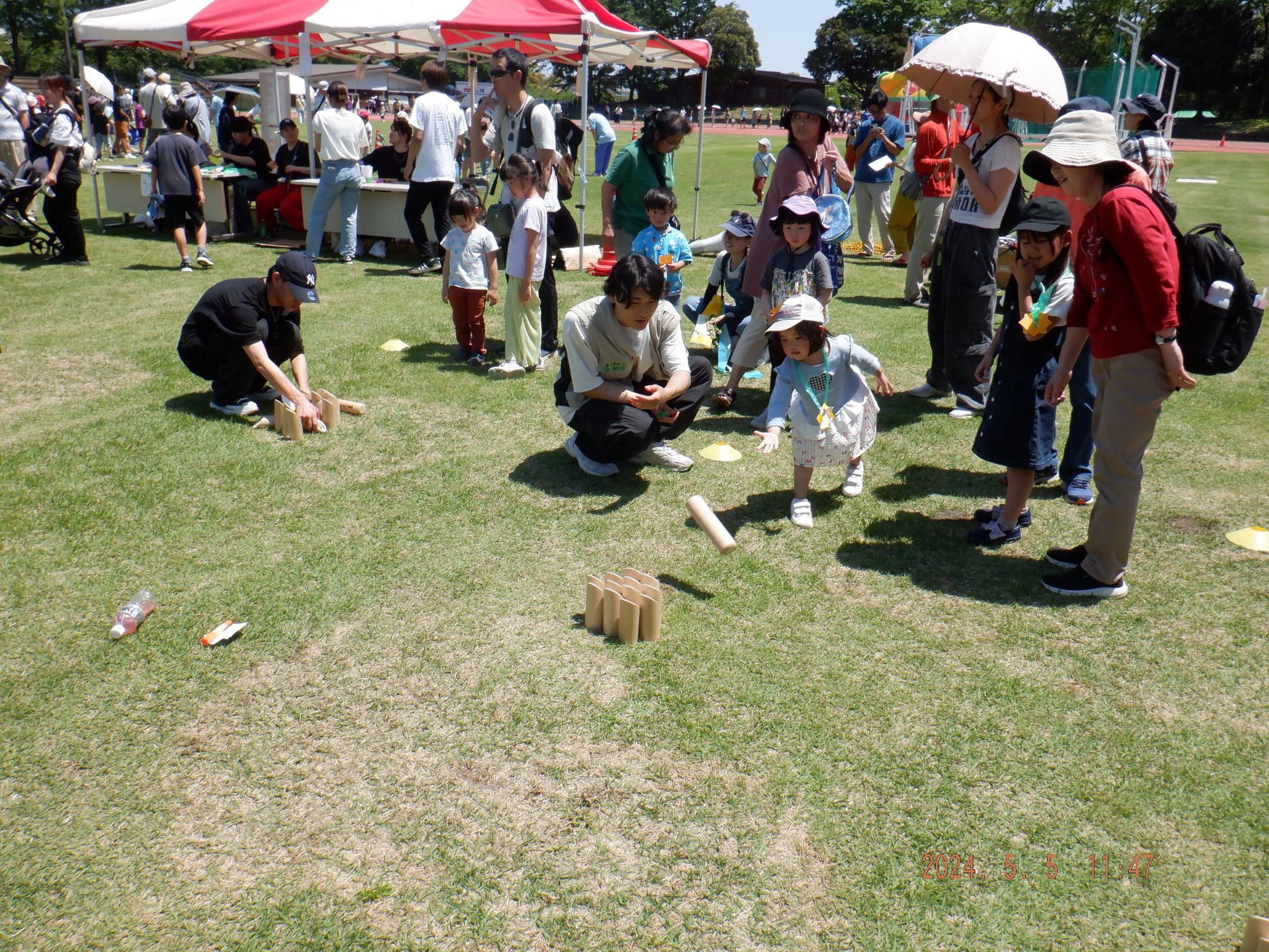 晴天の屋外イベント会場の芝生の上で、数本の木製ブロックを立てて倒すモルックのようなゲームを、しゃがみこんで見守る大人と、その周囲に集まり楽しんでいる複数の子どもたちの様子を写した写真