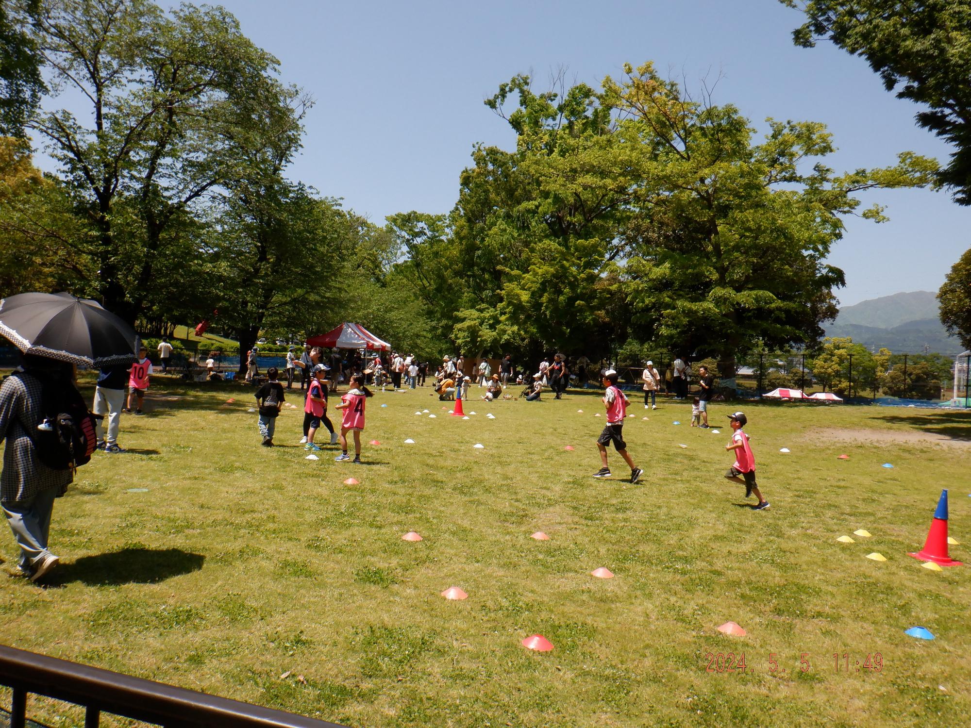 晴れた公園の芝生広場で、ピンク色のコーンや目印が置かれたエリアを、数人の子どもたちが運動着で走り回ったり歩いたりして楽しんでおり、その周囲で多くの人々がイベントに参加している様子の写真