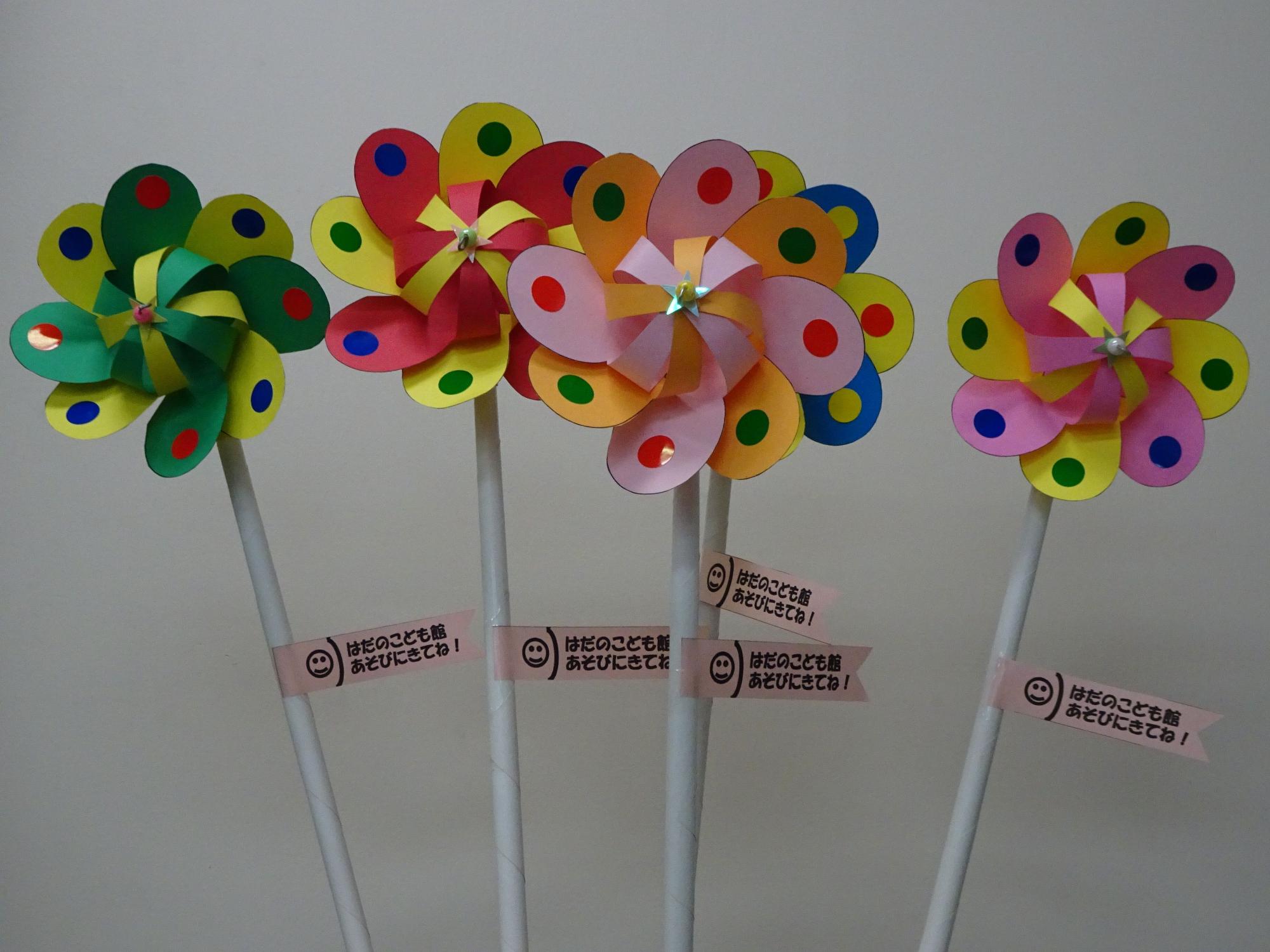白っぽい壁の前で、花びらのようなカラフルな飾りがついた四つの風車が、白い棒に「はだののこども館あそびにきてね!」と書かれた旗とともに立てられている写真