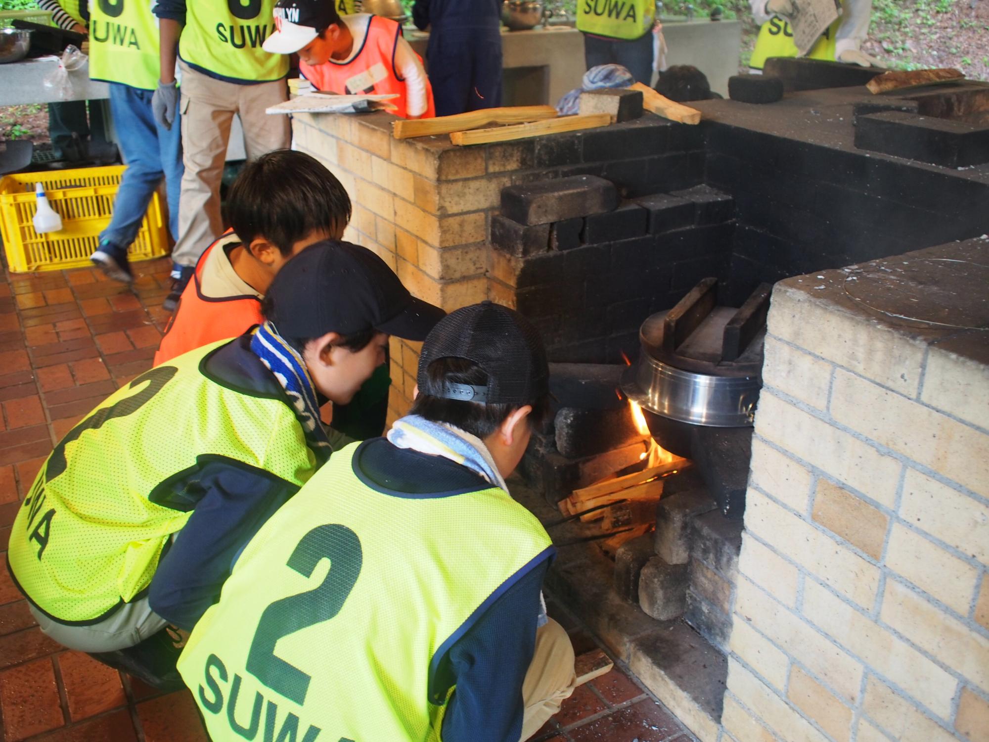 屋外のかまどの前で、黄色のビブスを着用した子供達がしゃがんで薪をくべている写真