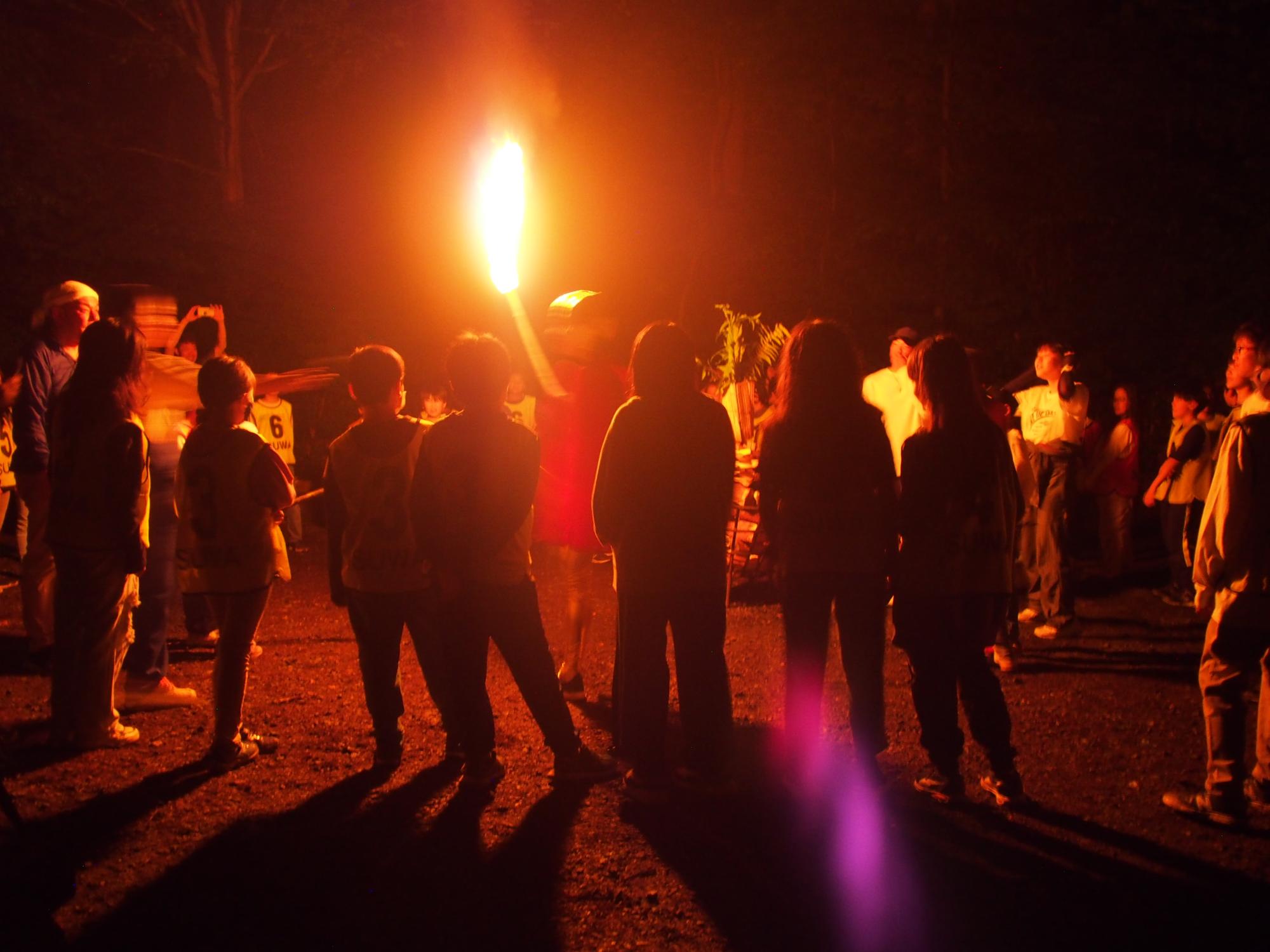 暗闇の中、複数の子供達と大人が焚き火を囲み、キャンプファイヤーを楽しんでいる写真