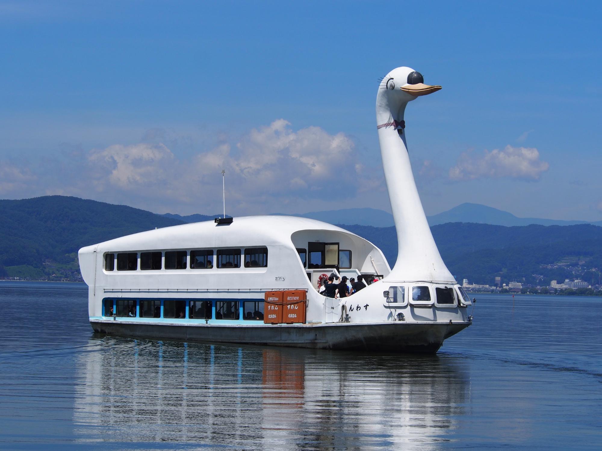 青空の下、白鳥の遊覧船が湖面を航行しており、乗客が外の風景を眺めている写真