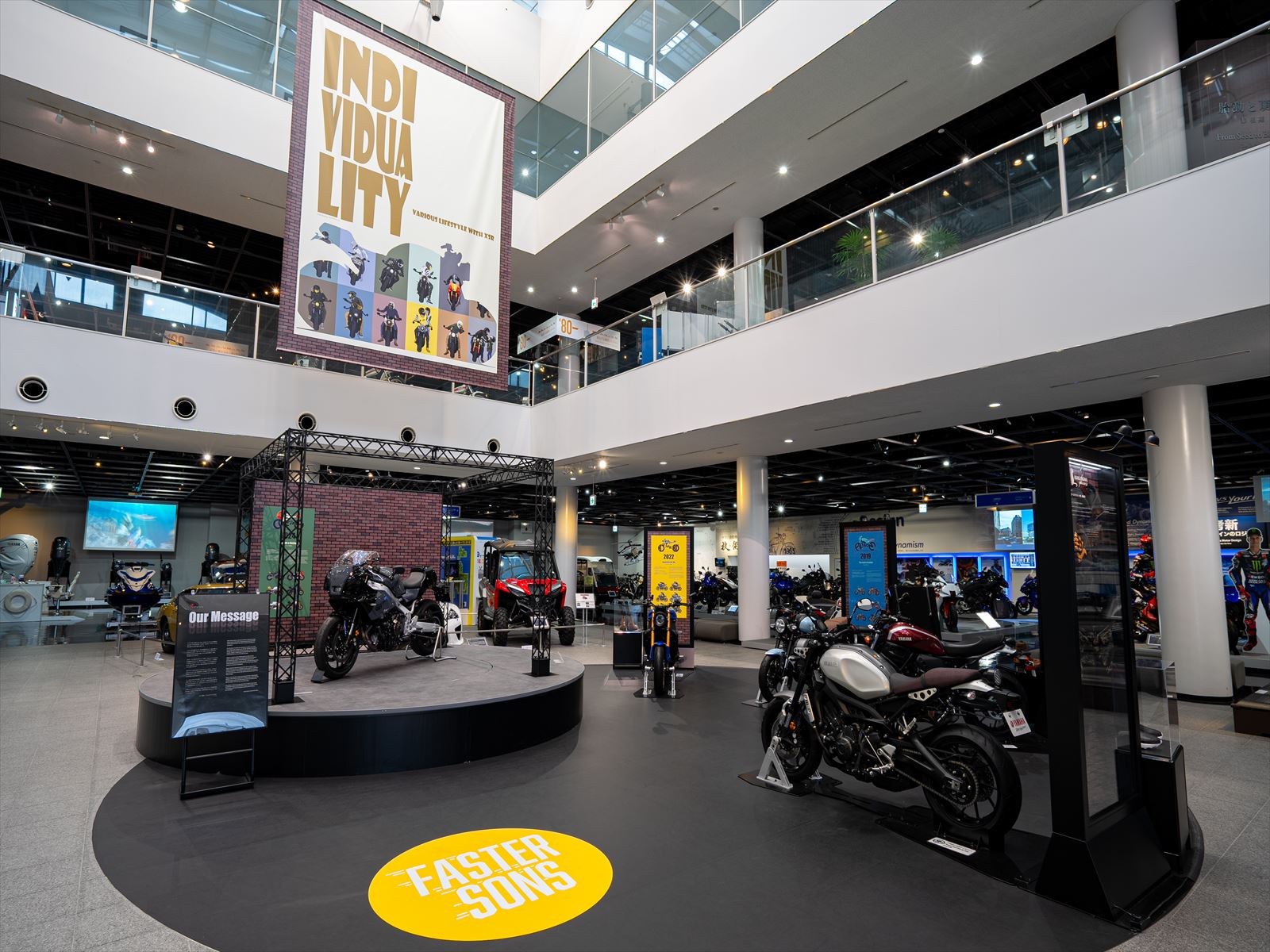 吹き抜けの広々とした展示スペースに、「INDIVIDUALITY」と書かれた大きな横断幕が吊るされており、様々な種類のバイクが展示されている写真