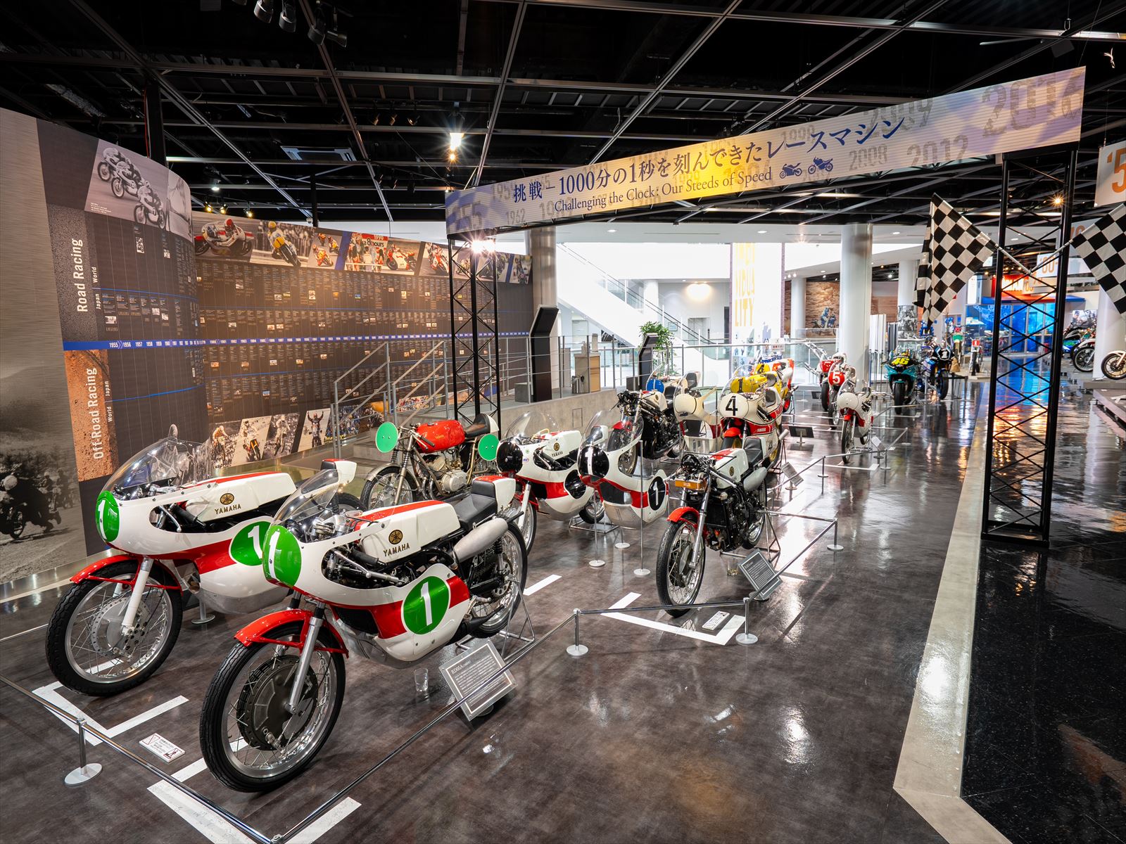 「挑戦 - 1000分の1秒を刻んできたレースマシン」と書かれた横断幕の下に、ゼッケンを付けたクラシックなレーシングバイクがサーキット場のように展示されている写真