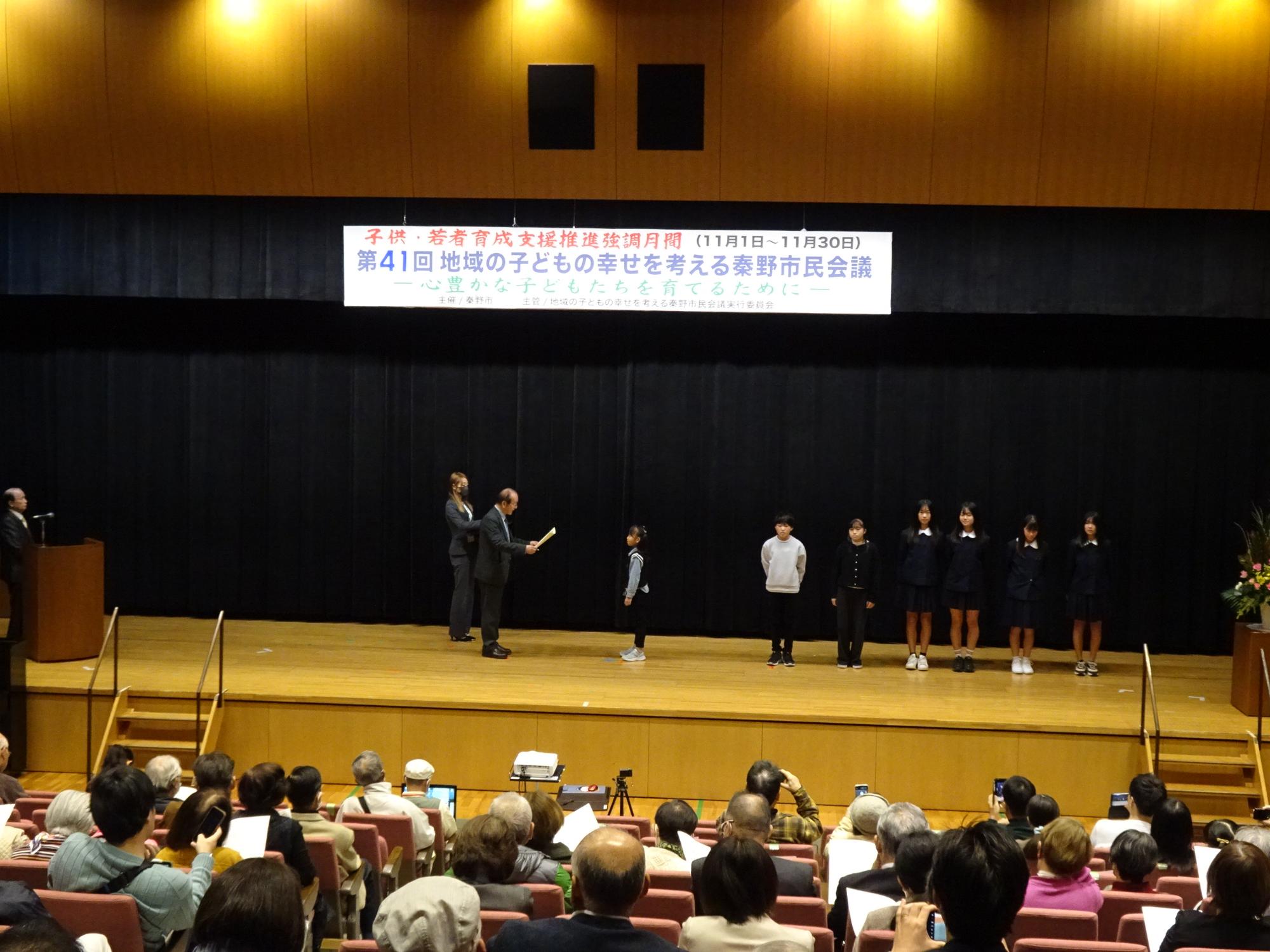 講堂のステージ上で、黒い幕を背景に「子供・若者育成支援推進月間 第41回地域の子どもの幸せを考える秦野市民会議」の横断幕の下、大人2名と中央で表彰を受ける女子児童、そして整列した制服姿の生徒たちを、客席側から写した写真
