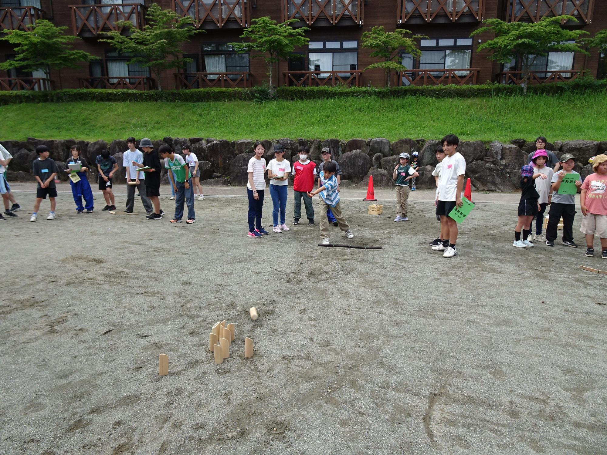 表丹沢野外活動センターの広場で、木製の棒を使って木製のピンを倒そうとしている子供とそれを見守る子供達の写真