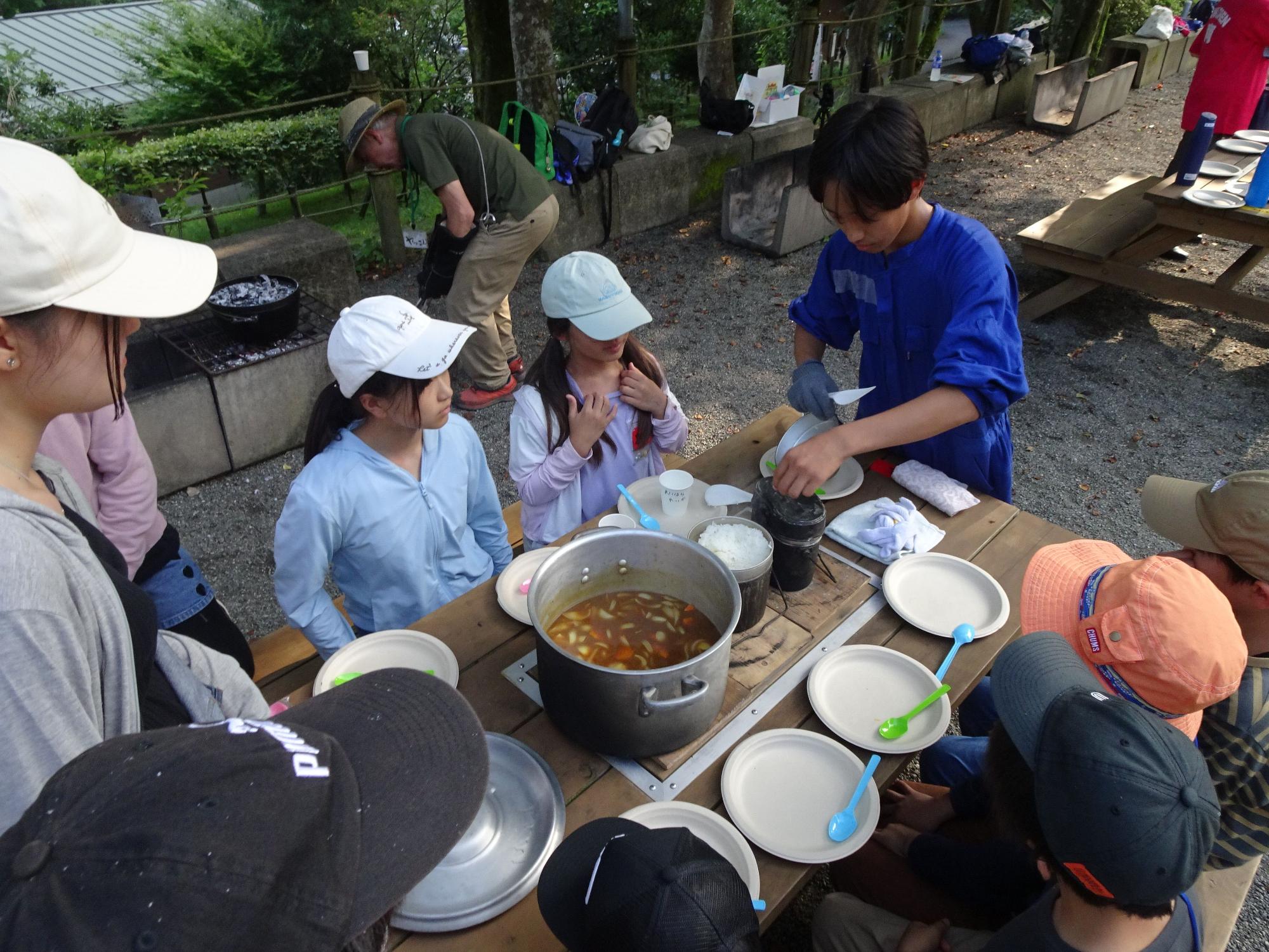 大なべで作ったカレーとお皿が並べられた屋外のテーブルに子供達と大人が座り、青いシャツを着た男の子が飯盒で炊いたご飯を開けようとしている写真