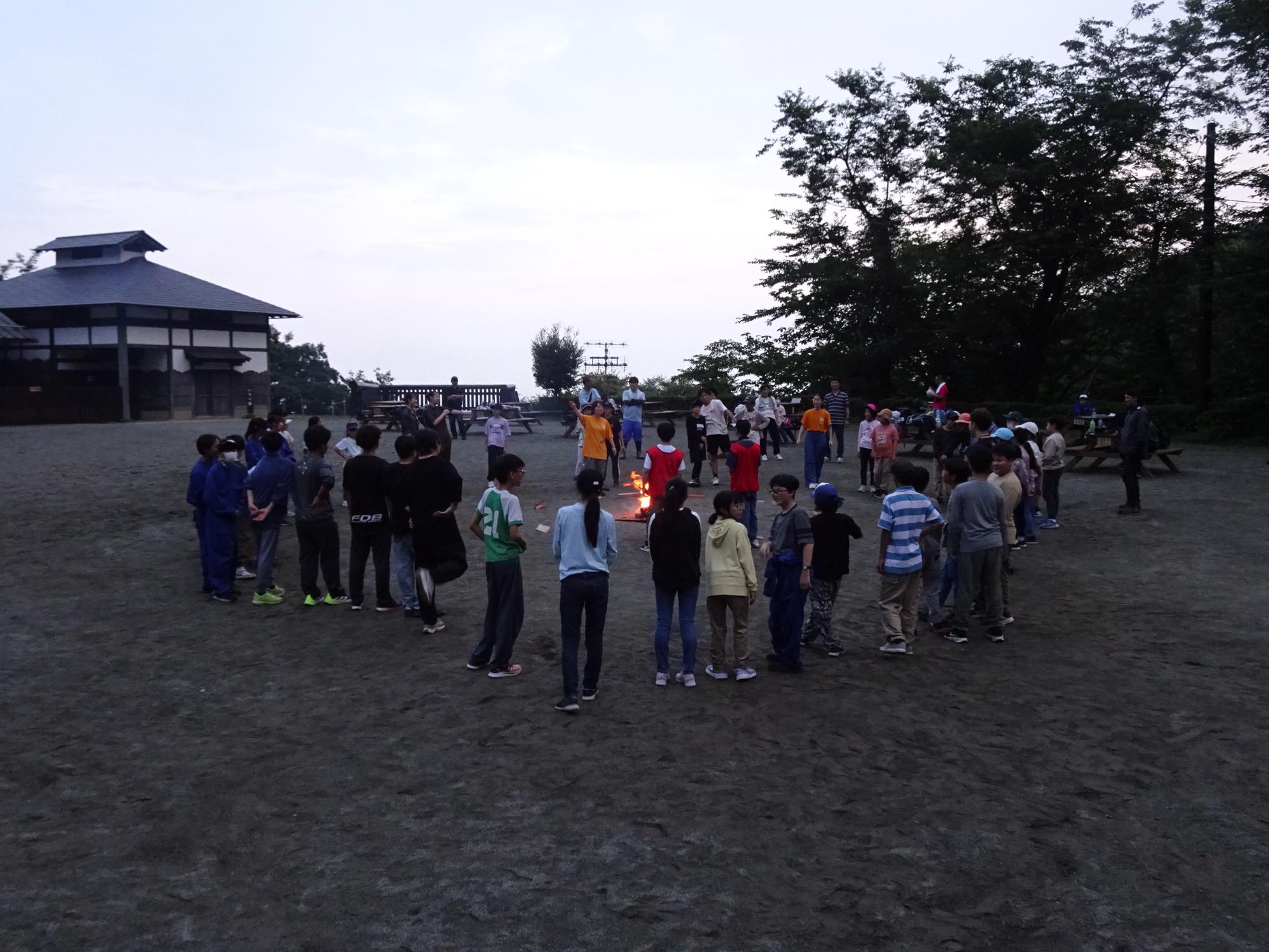 薄暗い外で、複数の子供達と大人が焚き火を囲んでキャンプファイヤーを楽しんでいる写真