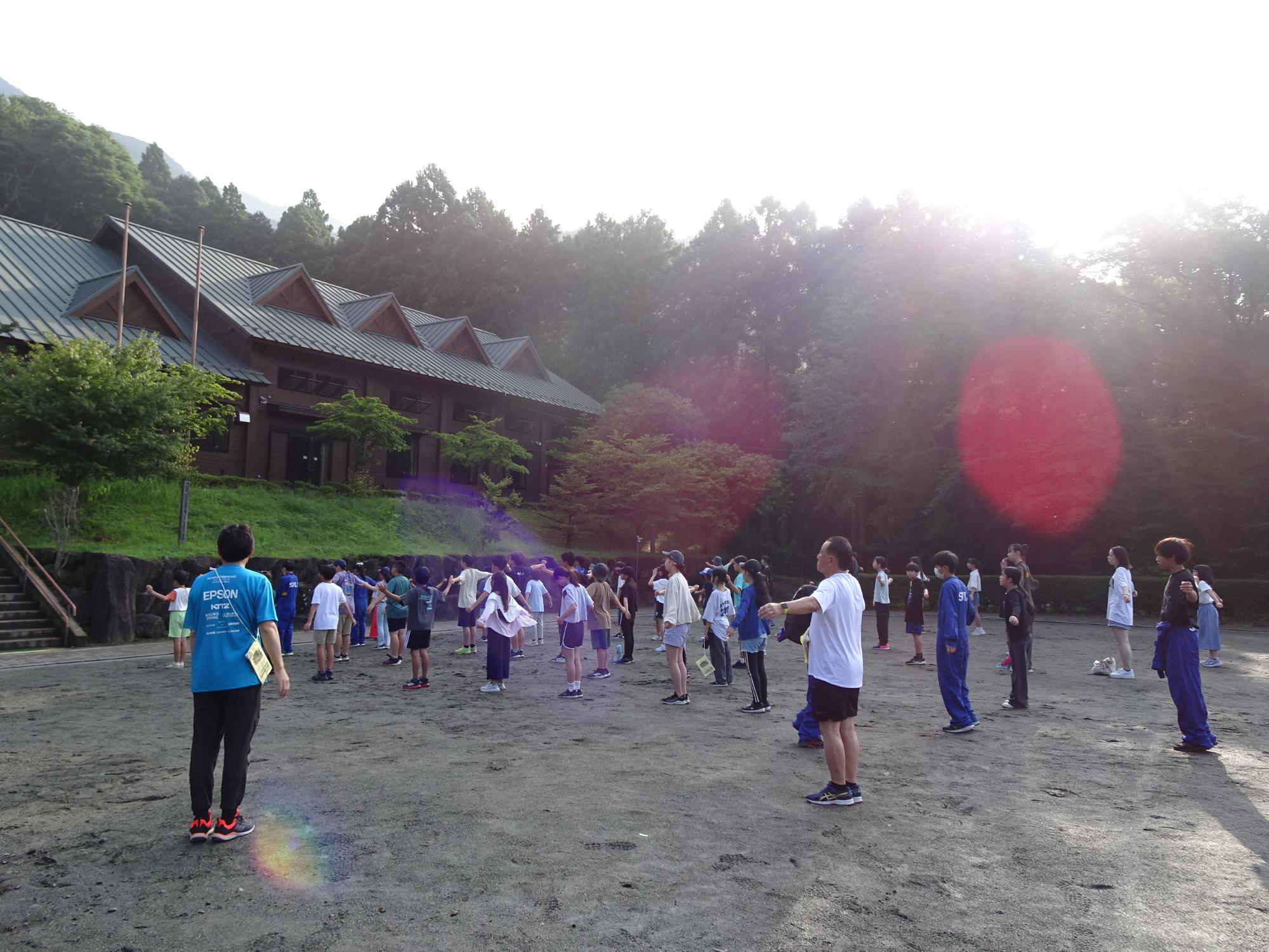 自然に囲まれた施設前の広場で、複数の子供達と大人が、腕を広げてラジオ体操を行っている写真