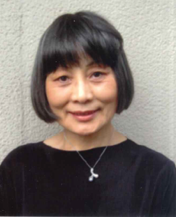 黒い服を着て、短めの黒髪に明るい色のハイライトが入った大畑　眞由美 氏が、薄いグレーの壁を背景に、わずかに口を開けて優しく微笑んでいる様子のポートレート写真