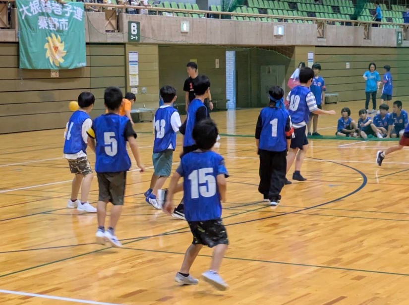 体育館の屋内コートで、青いゼッケンを着用した小学生くらいの子供たちがスポーツをしている様子を、観客席側から捉えており、背景には「大根巻寄子連 奉野市」と書かれた緑色の横断幕が見えるスポーツ写真