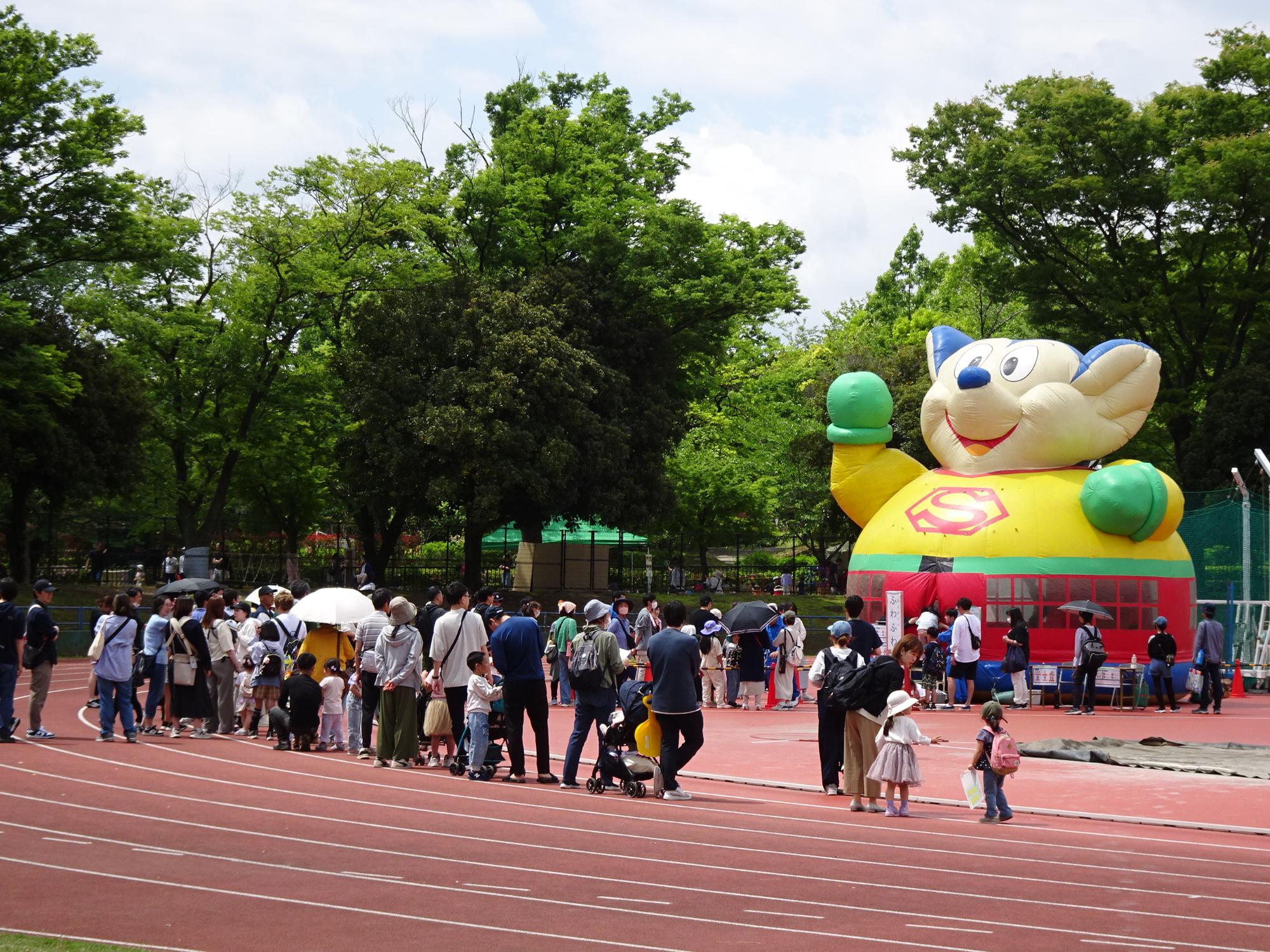 青空の屋外イベント会場で、赤く舗装されたトラックの向こうに、両手を挙げて歓迎しているような巨大な犬のキャラクターのエア遊具があり、その周りに多くの親子連れや参加者が集まっているイベント風景の写真