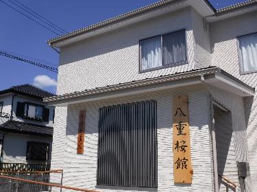 白くて細かいレンガ調の外壁が特徴的な二階建ての建物で、正面には黒い縦格子の大きな窓、右側には「八重桜館」の縦書きの木製看板、左側には「千村児童室」の小さな木製看板が掲げられた千村児童室外観写真