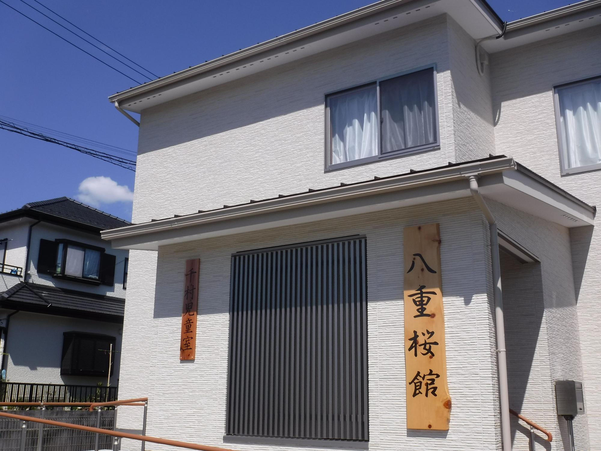 白くて細かいレンガ調の外壁が特徴的な二階建ての建物で、正面には黒い縦格子の大きな窓、右側には「八重桜館」の縦書きの木製看板、左側には「千村児童室」の小さな木製看板が掲げられた千村児童室外観写真