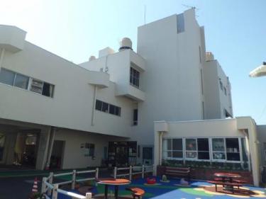 建物の手前には、子供の遊び場らしきカラフルな青いマットの地面、小さな赤いテーブルとベンチが並び、白い柵で囲まれているはだのこども館の外観写真