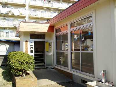 クリーム色の壁に赤い屋根の平屋の建物で、L字になった入口の横には大きな窓があり、手前には植え込みと屋外の流し台がある広畑児童館外観写真