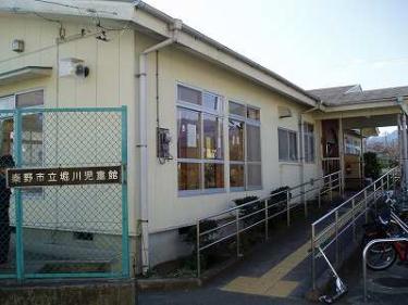 薄い黄色の外壁の平屋建ての建物で、建物の入口横には手すり付きのスロープが設けられており、手前の緑色の金網には「秦野市立堀川児童館」と書かれた看板が取り付けられている堀川児童館外観写真