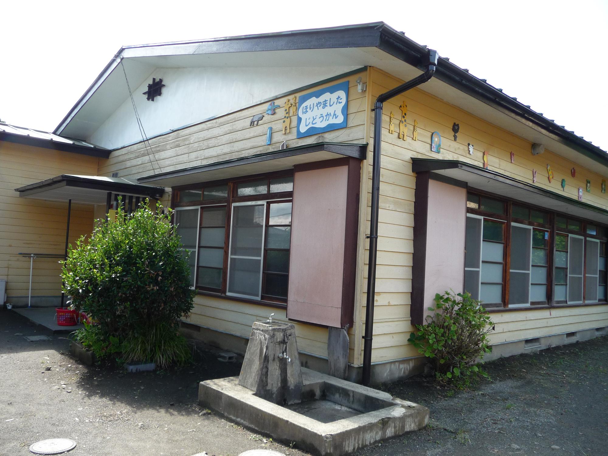 手前の壁に8つの窓が並んでいる茶色の板張りの屋根が白い平屋建ての建物で、左奥には小さな別棟がある堀山下児童館外観写真