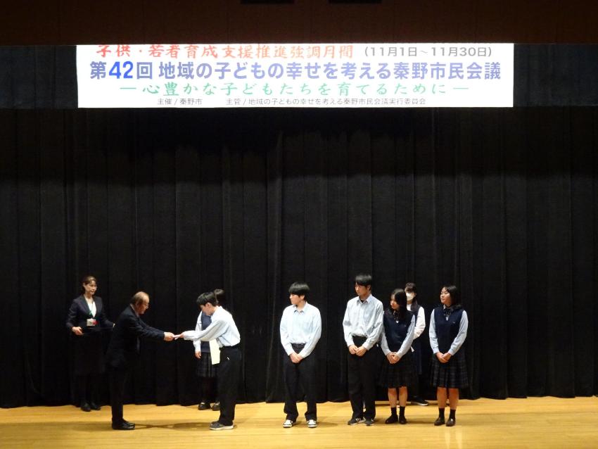 講堂のステージ上で、黒い幕を背景に「子供・若者育成支援推進月間 第42回地域の子どもの幸せを考える秦野市民会議」の横断幕の下、大人2名と中央で表彰を受ける女子児童、そして整列した制服姿の生徒たちを、客席側から写した写真