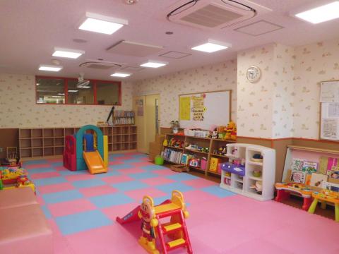 ピンクと水色のマットが敷かれた床に、カラフルな室内用の滑り台や本棚、おもちゃが置かれており、小さな子供達が自由に遊べる室内スペースの写真