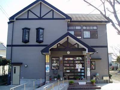 灰色で統一された外壁に焦げ茶色の装飾が施された洋風の二階建ての建物で、玄関には傾斜のついた屋根があり、正面の窓ガラスに多数のポスターが貼られた玄関からスロープが設置されている沼代児童館外観写真