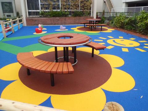 ひまわりと太陽のイラストが描かれたゴムチップ舗装の地面に、円形のテーブルと椅子が設置された屋外の遊び場の写真