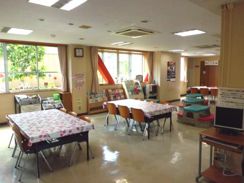 6人掛けの椅子とテーブルが複数設置され、壁際に本棚や絵本が置かれており自由に遊べる室内スペースの写真
