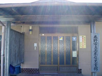 薄茶色の壁と焦げ茶色の屋根の建物で、玄関には金色の枠の入った曇りガラスのドアがあり、右の壁には「秦野市立三屋児童館」と書かれた木製の看板が立てられている三屋台児童館外観写真