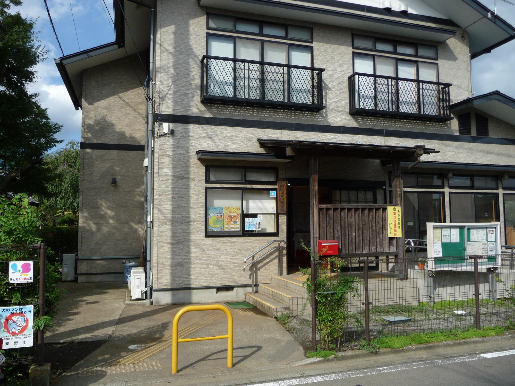 ベージュの外壁と白い切妻屋根の建物で、二階には黒い格子状の手すりが付いた窓が二つ、一階には木製の柵に囲まれた入り口と大きなガラス窓が並び、手前に緑色の掲示板が設置されている戸川児童館外観写真